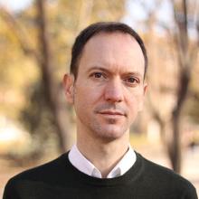 A person with short dark hair, wearing a white shirt under a black jumper.