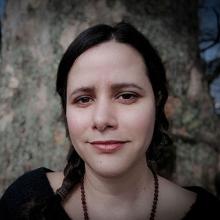 A person with dark hair, plaited, wearing a dark top and beaded necklace.