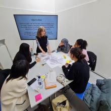 Gina Olsson standing in front of screen and behind a desk teaching a small group of students.