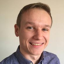 A person with short, light brown hair, wearing a blue shirt and smiling at the camera