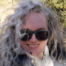 A person with curly silver hair, wearing sunglasses and smiling.