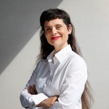 A person with long dark hair, wearing a white shirt and smiling at the camera.