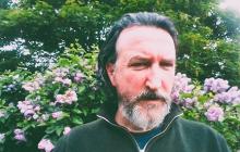 A person dark hair and a beard, standing in front of a flowering bush.