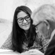A black and white picture of a person with dark hair, glasses, and smiling at another, older person.