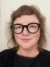 A person with short, light brown hair, wearing black rimmed glasses and smiling at the camera.
