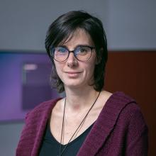 A person with short dark hair, wearing glasses, smiles to the camera.