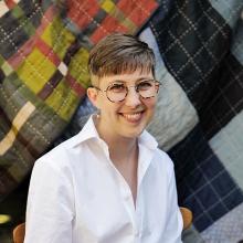 A person with short light brown hair, wearing glasses and a white shirt, sitting against a background of multi-coloured quilted blankets.