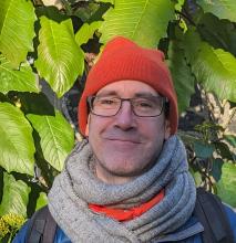 A person wearing glasses, an orange hat and a grey scarf stands in front of a leafy tree.