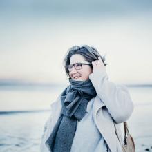 A person wearing a coat and big grey scarf, looking towards the sea