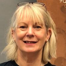 A person with short blond hair, wearing glasses on their head and smiling at the camera.