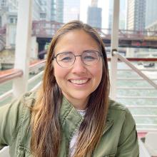 A person with long brown hair, wearing glasses and a green jacket, smiling at the camera.