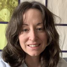 A person with long dark hair, wearing a white top and smiling to the camera.