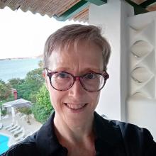 A person with short, blond hair, wearing glasses and a dark shirt, with a view of an outdoor pool, trees and the sea in the background.