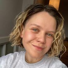 A person with short, blond curly hair, wearing a grey tshirt and smiling to camera.
