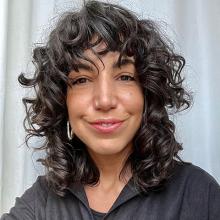 A person with dark curly hair, wearing a black shirt and smiling at the camera.