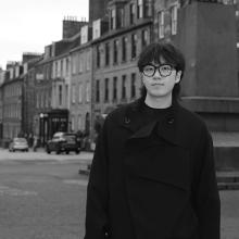 A black and white image of a person with short dark hair, wearing black glasses, and a black coat. They are standing on Castle Street in Edinburgh.