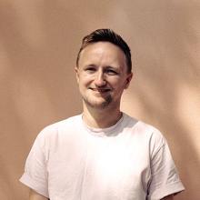 A person in a white t-shirt, standing against a light bisque coloured wall. The sunlight filters through the branches of a tree on to them.