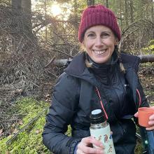 Annie Gallagher in a maroon hat, black coat, sitting in a forest and holding a flask and cup. Annie is smiling at the camera.