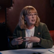A person with shoulder length strawberry blonde hair, wearing glasses, a suit jacket and white shirt. The person is in the middle of saying something and gesticulating with their hands.