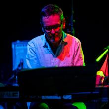 Musician Chris Letcher playing the keyboard at a gig. They are cast in blue, pink and green stage lights.