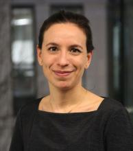 Larissa has their hair tied back and is wearing a black top with a square neckline. They are looking into the camera and smiling.