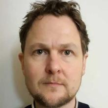 A person with short light brown hair, wearing a black jumper. They're looking directly to the camera.