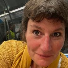 A person with short, dark brown hair, wearing a yellow knitted top, looks to the camera.