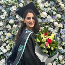 A person wearing graduation robes, standing in front of a floral wall, holding a bunch of flowers and smiling to the camera.