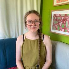 A person with long dark blonde hair, tied in a plait, sits on a sofa in front of a green wall which has framed pictures on it.