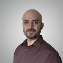 A bald person with a dark beard. They're half turned to the camera, wearing a maroon shirt.