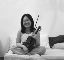 A black and white image of a person with shoulder length dark hair, holding a violin and smiling at the camera.