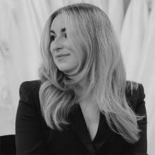 A black and white image of a person with long light coloured hair, looking sideways and smiling. They're wearing a dark coloured blazer.