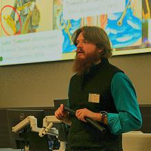 A person standing in front of a projector screen, talking to a class whilst holding an instrument that resembles a black pipe.