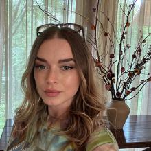 A person with long hair, looking into the camera. Behind them is a console table with dried flowers in them and sheer curtains behind that.