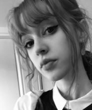 A black and white image of a person with a fringe, black winged eyeliner. They're looking down at the camera.