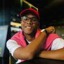 A person wearing a salmon pink vest, white tshirt and hat. They're smiling to the camera.