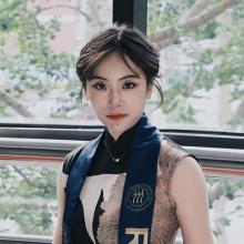 A person stands in front of a window looking out onto trees. They wear a dress and a blue sash draped around their neck. They're looking directly to camera.