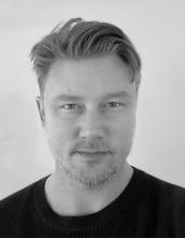 A black and white photo of a person with short light hair and they're wearing a black tshirt. They're looking directly into the camera.