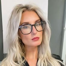 A person with long, blonde/silver hair, looking at the camera. They wear big, black glasses and are standing in front of a wall with a piece of white paper on it.