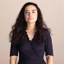 A person with long dark, wavy hair, wearing a navy blue, scalloped edge wrap dress. They look directly into the camera.
