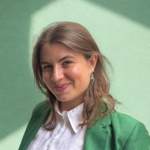A person with light brown coloured hair, smiles at the camera. They are wearing a white shirt and green jacket, and they're standing against a green background.