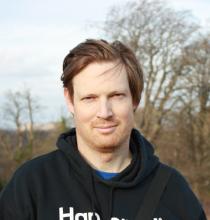 Dr Tom Mudd stands outside in a black hooded jumper. There are trees in the background.