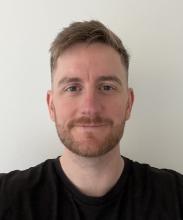 A person with sandy coloured hair, smiles to the camera. They're wearing a black tshirt and stand in front of a white wall.