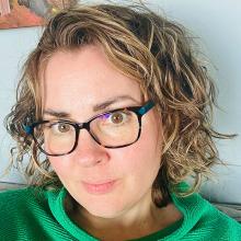 A person with short, curly brown/blond hair, wearing glasses and a green top, smiles to the camera.