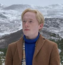 A person standing against a backdrop of snowy mountains. They wear a blue jumper and tan coat.