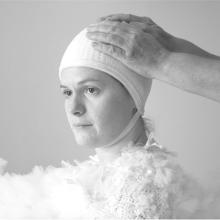 A black and white image of a person with a white cap on, and layers of ballet tulle wrapped around them. A person's hands are resting on their head.