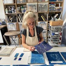 Susan Cross sitting at a table in a studio, with cyanotype prints of plants.