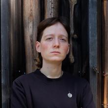 A person with short hair, wearing a navy blue jumper, stands in front of a what looks like a wooden cabin wall.
