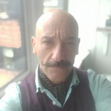 A person with a shaved head, a moustache twirled up at the ends, looking straight to the camera.