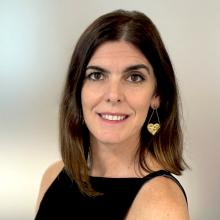 Pilar smiles to the camera. Their hair is dark brown and shoulder length and they wear a black strappy top and gold heard earrings.